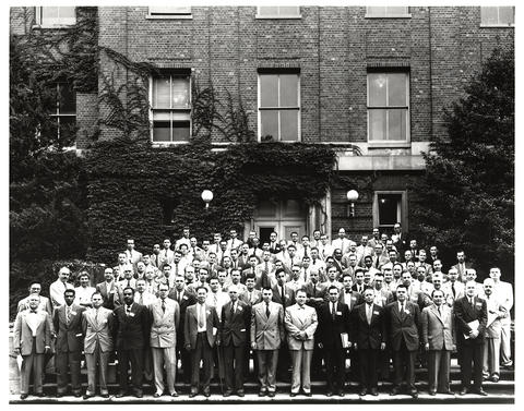 Men pose outside a brick building in a historical black-and-white photo.