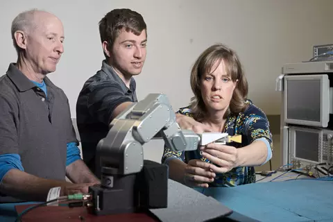 Two men and a woman stand at a table holding a robotic arm. 