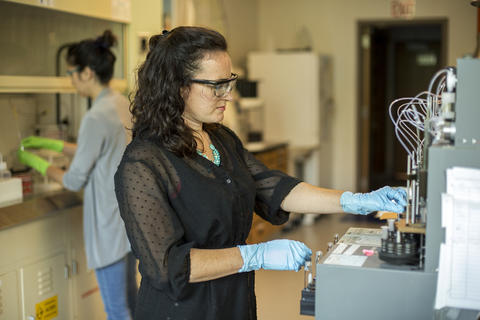A woman with safety goggles and gloves stands at a lab bench.