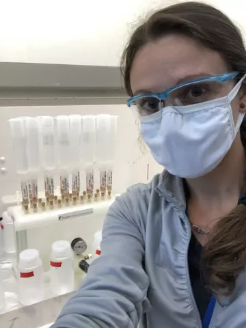 A woman wearing safety goggles and a mask poses in front of a cabinet holding vials.