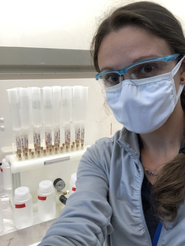 A woman wearing safety goggles and a mask poses in front of a cabinet holding vials.