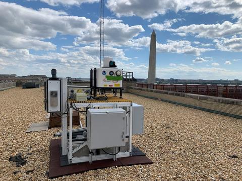 Gray metal scientific instrument is mounted on flat roof of building with Washington Monument in background.