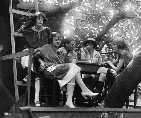 Four women and a man sitting at a table in a tree house.  The man seems to be talking to two of the women