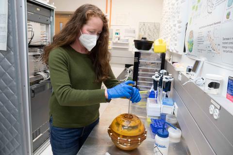 A woman wearing a mask stands over a yellow sphere at a lab bench. 