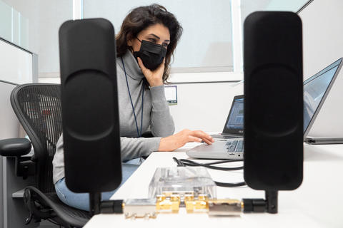 A woman wearing a face mask works at a laptop with communications equipment on the desk. 