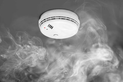 Smoke swirls around a white smoke detector on a ceiling. 