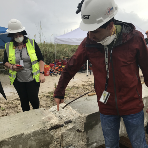 A man in a construction hat points to damage on a concrete column lying on the ground while another researcher looks on. 