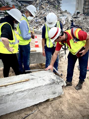 Four workers in hard hats look down at a damaged concrete pillar. 