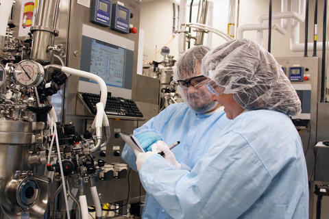 Two people in coveralls and hair covers check the readouts on laboratory equipment.