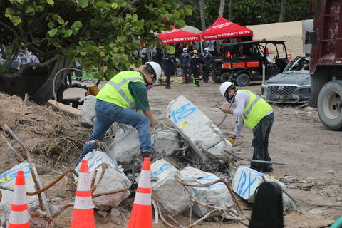 Two people in hard hats and other safety gear bend over broken concrete columns marked with blue spray paint, with tents and construction equipment in the background. 