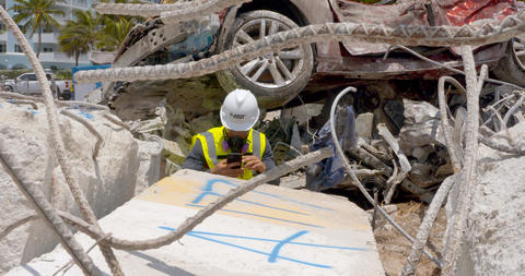 A person in a hard hat surrounded by debris including a crushed car takes a picture of a concrete block with a phone.