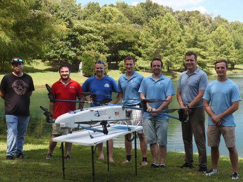 A group of seven people standing outside by a small body of water and several trees. The people stand behind a drone sitting on a table.