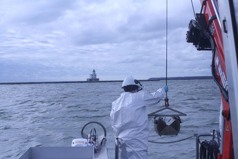 A researcher stands on a boat reaching for a cable holding a grabber device. 