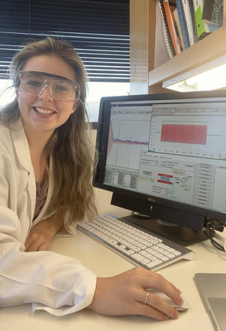 young woman in lab coat and safety glasses at a computer, she is smiling.