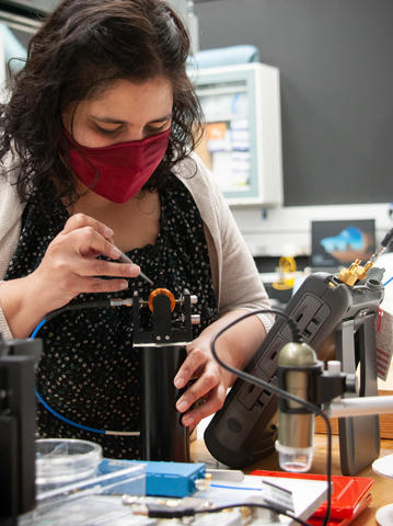 A woman wearing a face covering leans over a piece of scientific equipment, holding tweezers. 