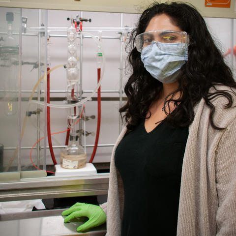 A woman wearing safety glasses and a face mask poses with chemistry equipment in the background.