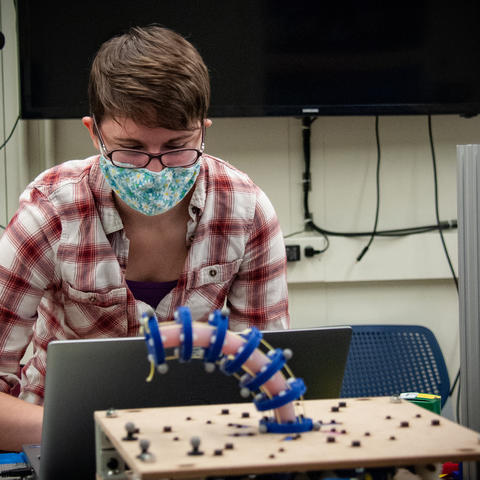 A woman in a face mask works at a laptop behind a curving prototype robot arm.