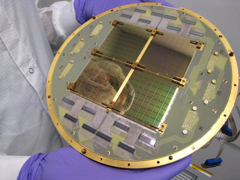 A person wearing purple gloves holds a circular reflective plate with square electronic chips at the center.