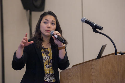 A woman (Danielle Santos) stands at a podium holding a microphone and gesturing with her other hand. 