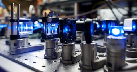 Lenses on short stands are lined up on a lab table with blue light shining through them. 