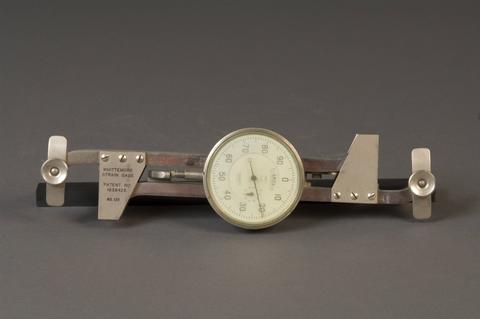 Metal scientific device with dial at center and attachment pieces at either end.