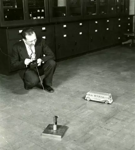 In a historical photo, a man crouches with a remote controller, looking at a toy-sized bus on the floor.