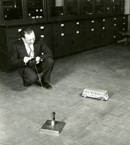 In a historical photo, a man crouches with a remote controller, looking at a toy-sized bus on the floor.