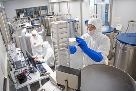 a large lab with people in white total body suits putting sample containers into huge vats of liquid nitrogen