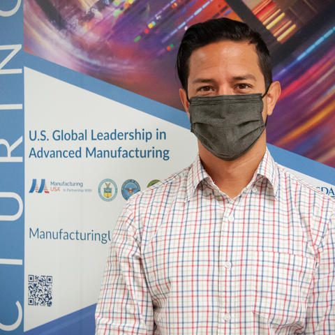 A man in a gray face mask poses in front of a Manufacturing USA poster. 