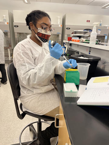 A woman (Diane Nelson) in a mask and lab coat works with plastic test tubes at a lab bench.