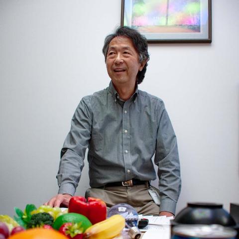 Yoshi Ohno poses behind a table holding brightly colored foods like red peppers and bananas. 