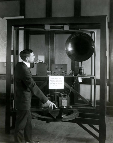 Historical photo shows a man manipulating a large device on a stand with large dial and handle at the bottom.