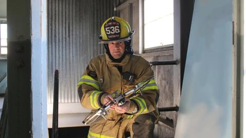 A man in firefighting gear stands inside a building holding a flat metal drone. 