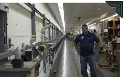 A man stands next to a long device running along the wall, used for calibrating tape measures.