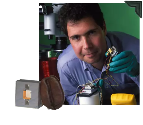 A dark-haired man holds an electronic device with wires connecting its parts; a computer chip with coffee bean for size is overlaid at lower left. 