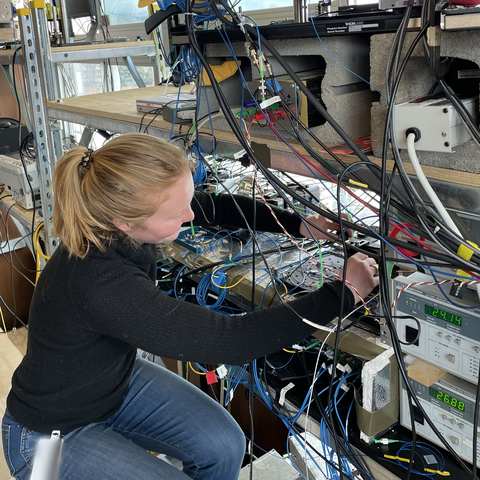 A woman with a ponytail sits next to a rack of electronics, adjusting wiring. 