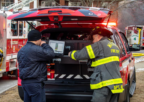 Two first responders stand at the open back of an emergency vehicle, looking at a computer screen, with firetrucks in the background.