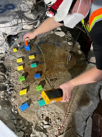 A researcher with a handheld device performs tests with small pieces of colored material lying on a large piece of concrete.