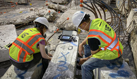 Two people wearing safety gear sit on either side of a concrete column lying on the floor, performing tests on it.