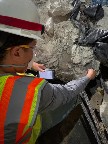 A researcher in safety gear holds a smartphone up to a large piece of concrete and rebar.