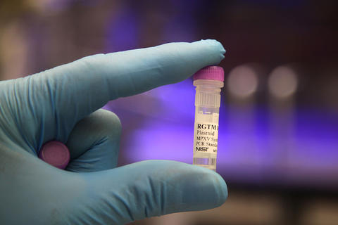 A gloved hand is holding up a small plastic vial labeled