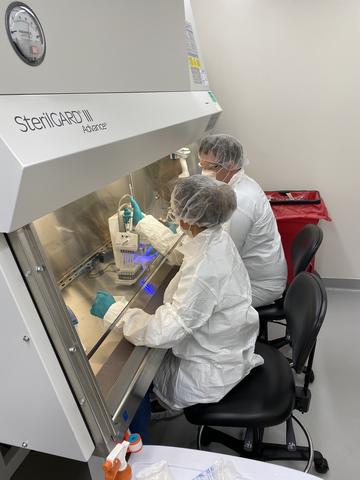 Two researchers in white coveralls and hair coverings use devices to fill vials on a metal counter inside a large transparent hood.