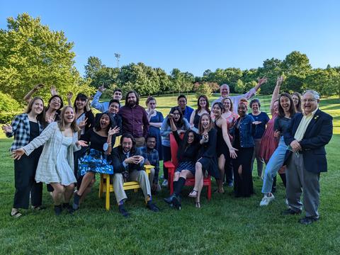 About 25 people pose for a group shot outdoors. Most are young, and many are laughing and waving. 