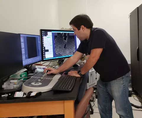 Thomas Cleveland stands at a desk, looking at a computer screen while turning a dial on a control panel. 
