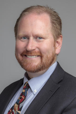 R. Austin Hicklin in a suit and tie. He has red hair, beard, and mustache, blue eye and a smile. 