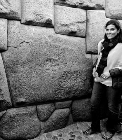 A woman poses smiling next to a big flat stone in a wall made mostly of smaller stones.