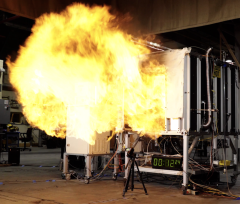 A fireball rushes out of the opening of a small metal compartment in a concrete room. 