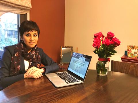 Yalda Saadat poses sitting at a table at home, next to an open laptop and a vase of roses. 
