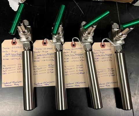 Photograph of four compressed gas sample cylinders with tags documenting composition. 