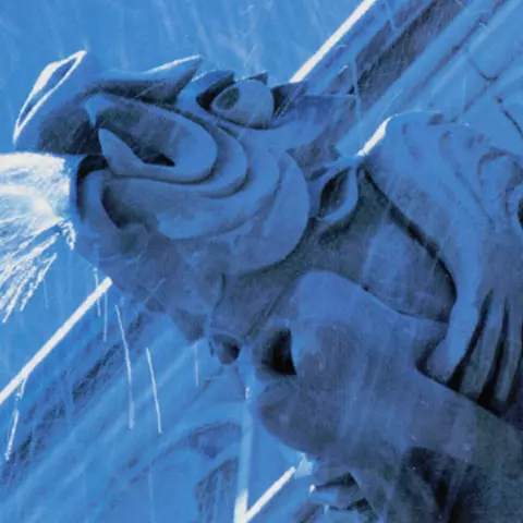 View from below of stone gargoyle spitting water and holding a large molar tooth. 